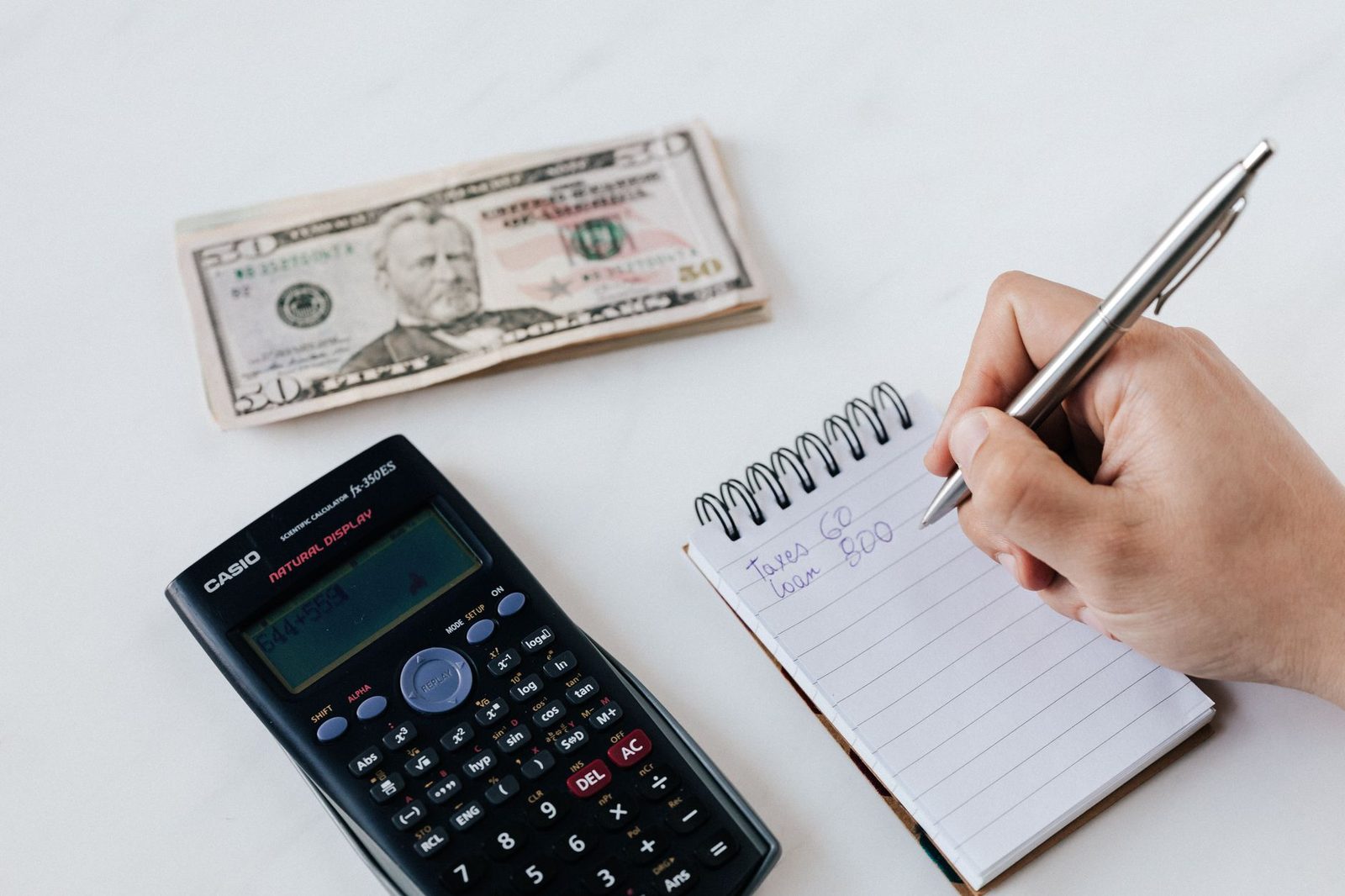 Overhead view of a hand writing in a notepad beside a calculator and a small stack of banknotes
