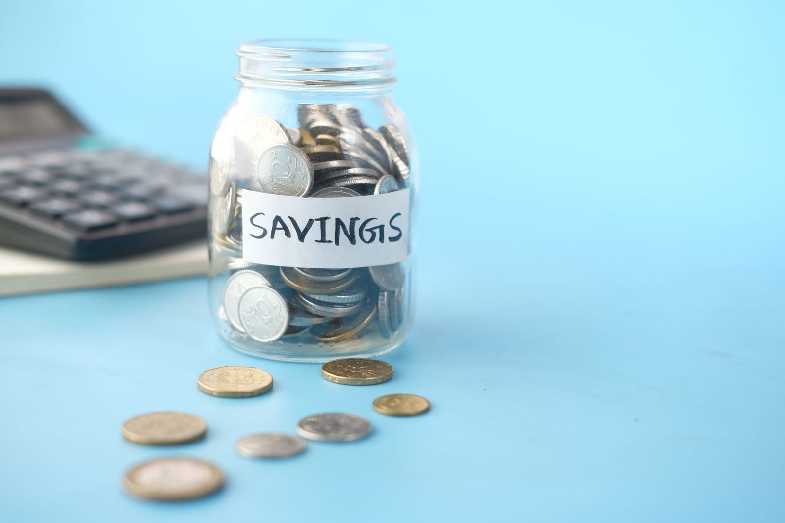 A glass jar labelled savings filled with coins, with a calculator behind it