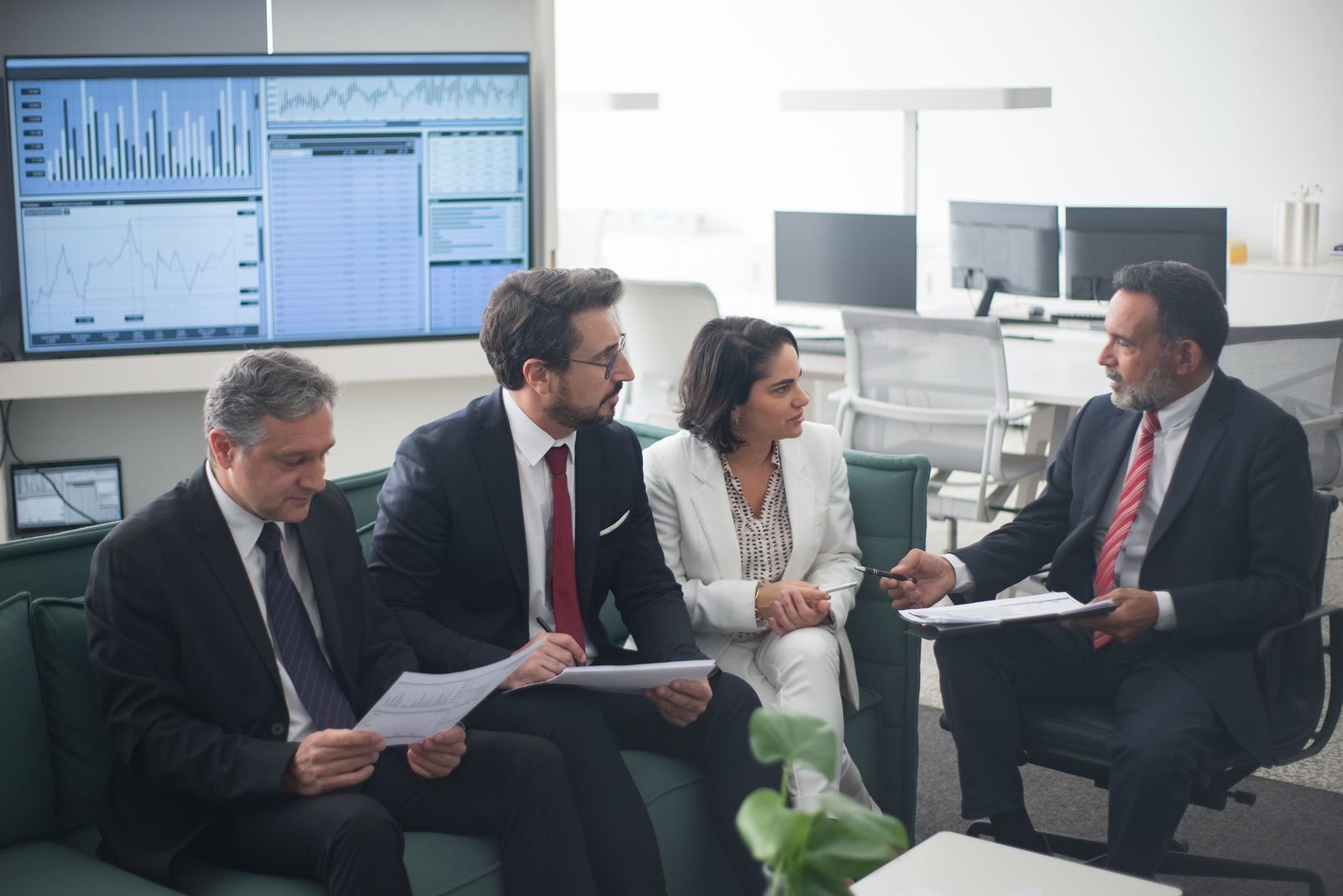 Colleagues meeting in an office with trading screens visible behind them
